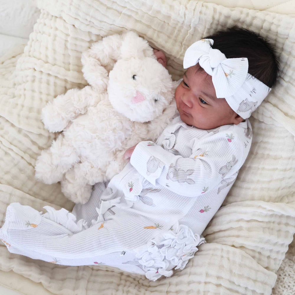 baby wearing hop little bunnies zipper one piece ruffle footie and matching large bow headwrap with cuddlebuddies bentley bunny stuffed toy
