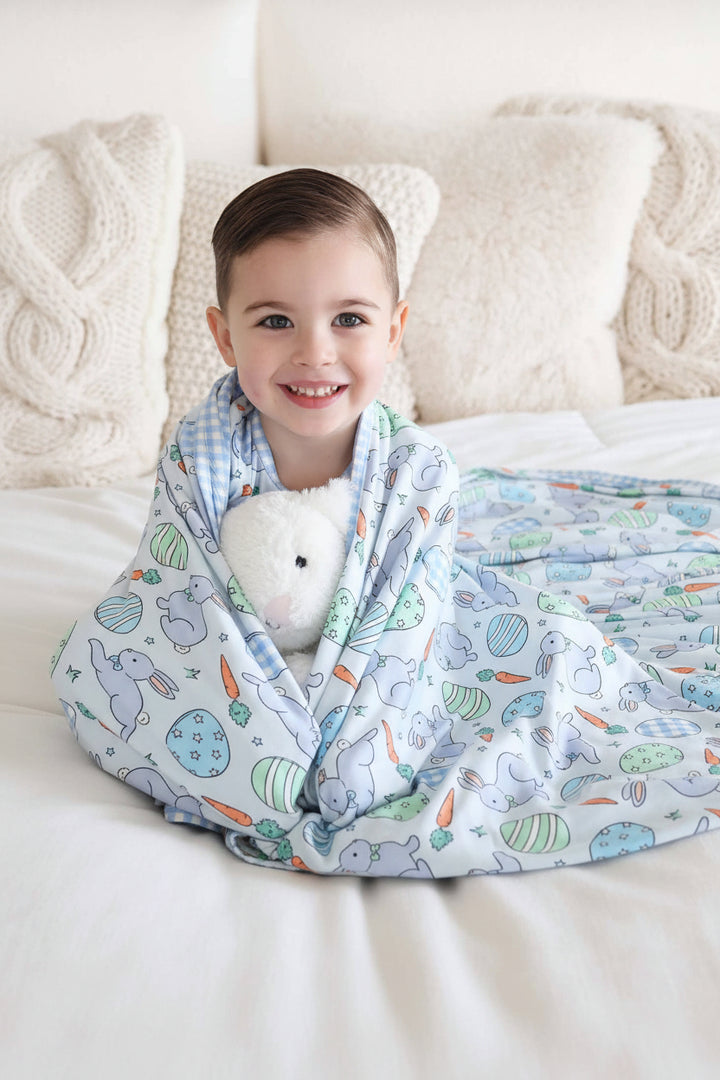 boy sitting on bed wrapped in blue hoppy spring double sided bamboo blanket holding a blue stuffed bunny 