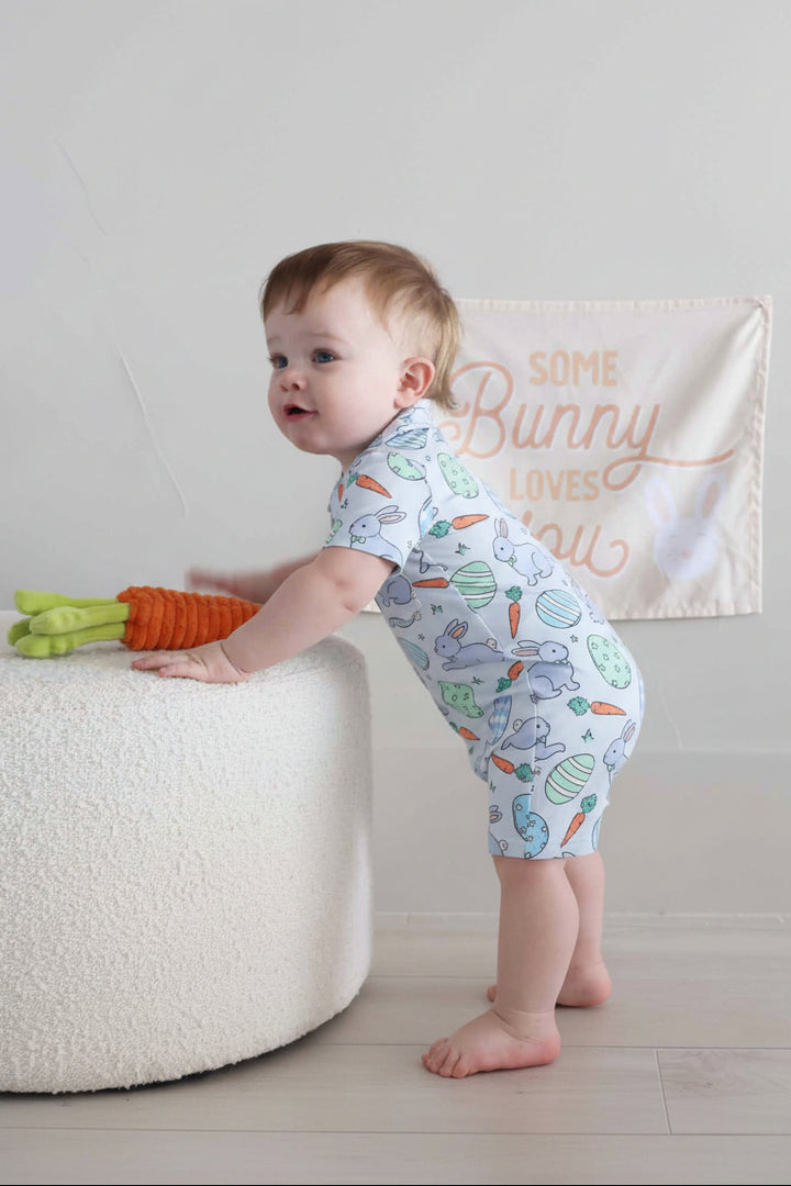 boy leaning over ottoman in blue hoppy spring short sleeve collared romper with a stuffed carrot 