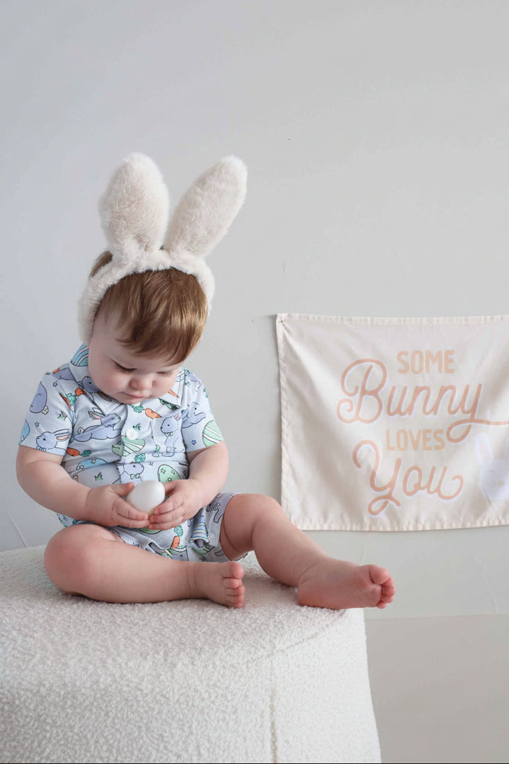 baby boy wearing the hoppy spring blue short sleeve easter front button romper with bunny ears in front of a some bunny loves you