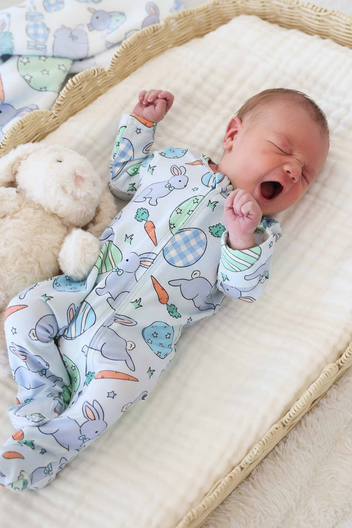 baby boy laying down wearing the hoppy spring blue zipper footie pajama with a white stuffed animal bunny next to him 