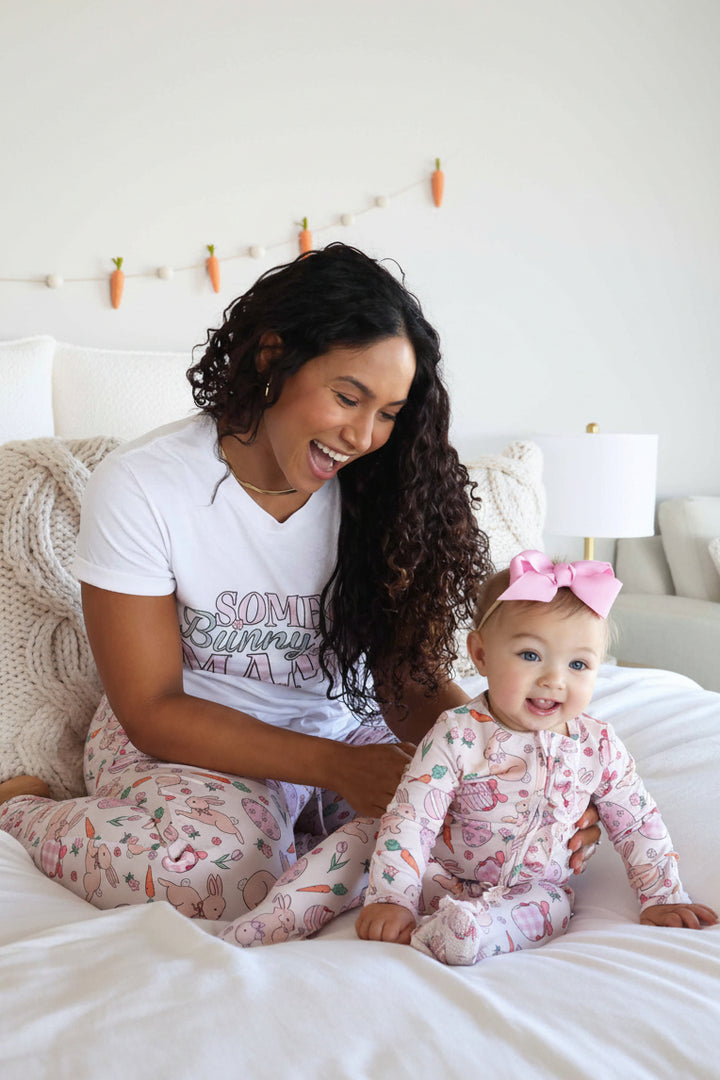 mom and baby sitting on bed wearing pink bunny, easter egg and carrot easter family matching pajamas 