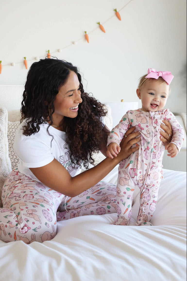 mom holding up baby girl standing on bed in hoppy spring pink ruffle zipper footie with a pink ribbon bow headband. mom is wearing matching pajama pants and a graphic tee