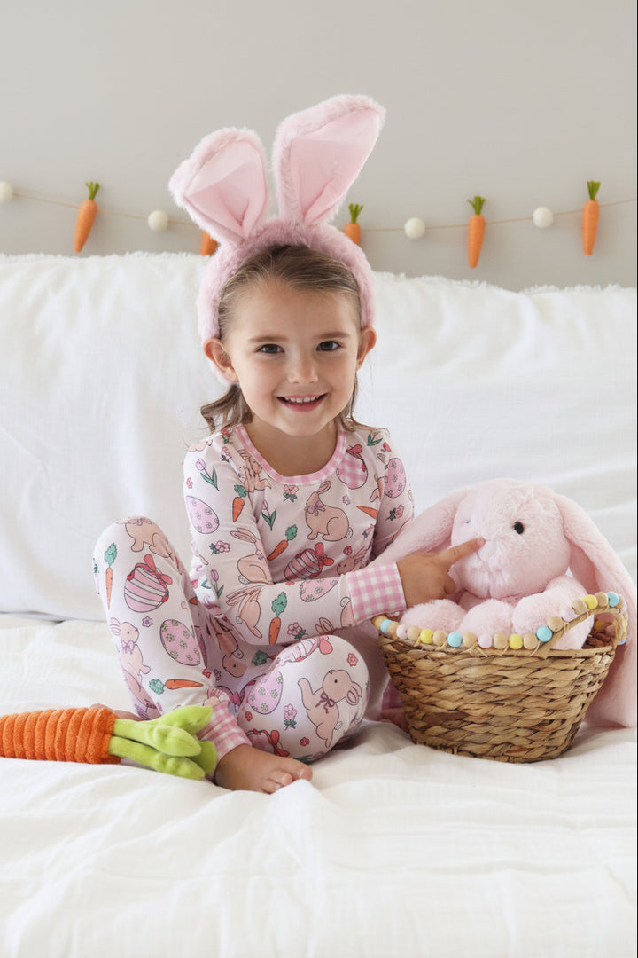 girl sitting on bed wearing hoppy spring two piece pajama set with bunny ears and pointing to a pink stuffed bunny in an easter basket 