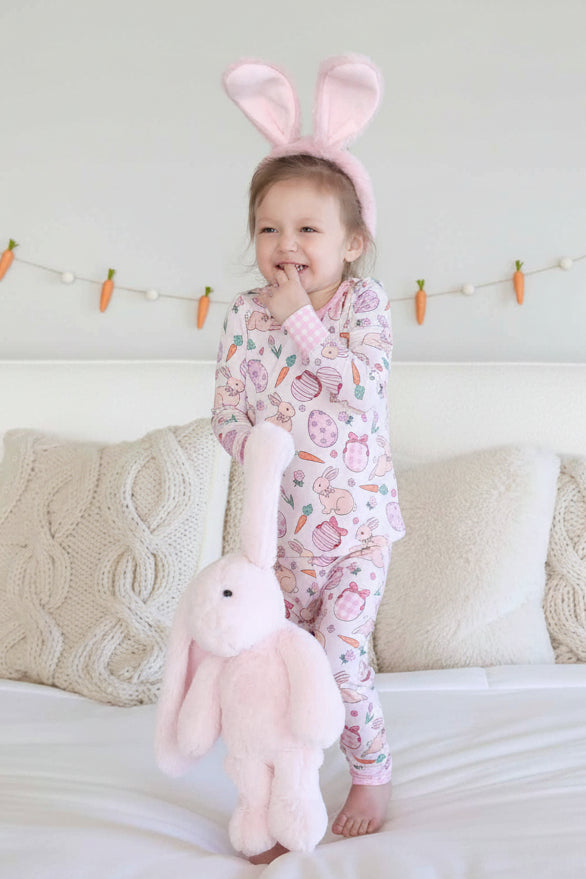 girl standing on bed in pink bunny, easter egg, carrot and flower two piece pajama set wearing pink bunny ears and holding a pink stuffed bunny 