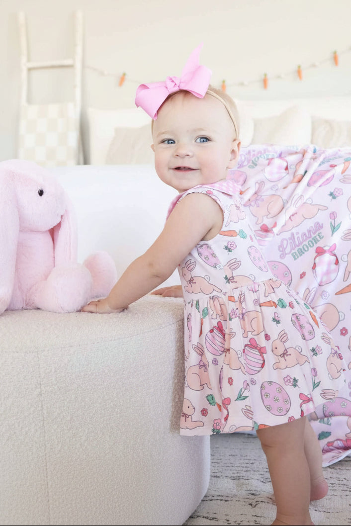 baby girl standing leaning on an ottoman wearing a pink easter themed bodysuit with a pink and white gingham peter pan collar 