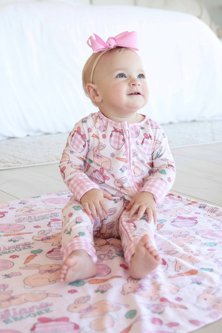 girl sitting on hoppy spring personalized blanket in pink wearing a matching convertible zip romper and a pink ribbon bow headband 