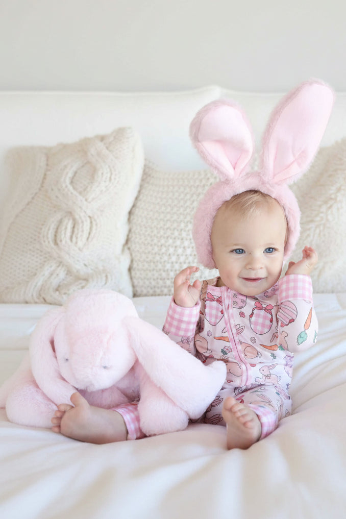 girl sitting on bed wearing hoppy spring pink convertible zip romper wearing bunny ears sitting next to a pink stuffed bunny
