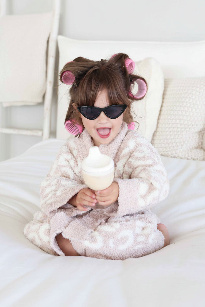 little girl with rollers in her hair and black sunglasses in a neutral leopard robe sitting on a bed 