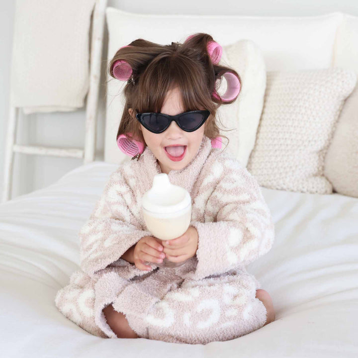 little girl with rollers in her hair and black sunglasses in a neutral leopard robe sitting on a bed 