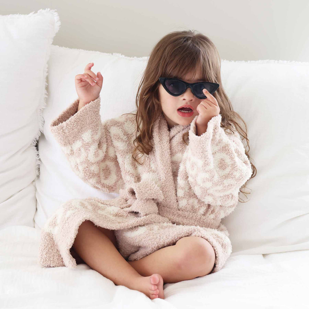 girl sitting on a bed wearing point black sunglasses wearing a beige and white leopard plush robe that ties at the waist 