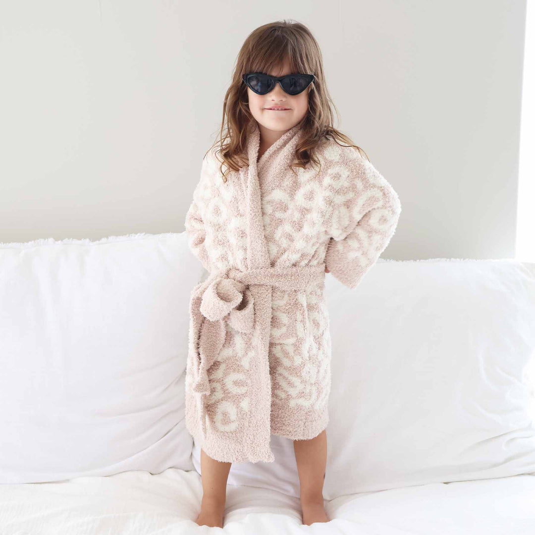 girl standing on a bed with her hands on her hips with black point sunglasses wearing a neutral leopard robe that ties at the waist 