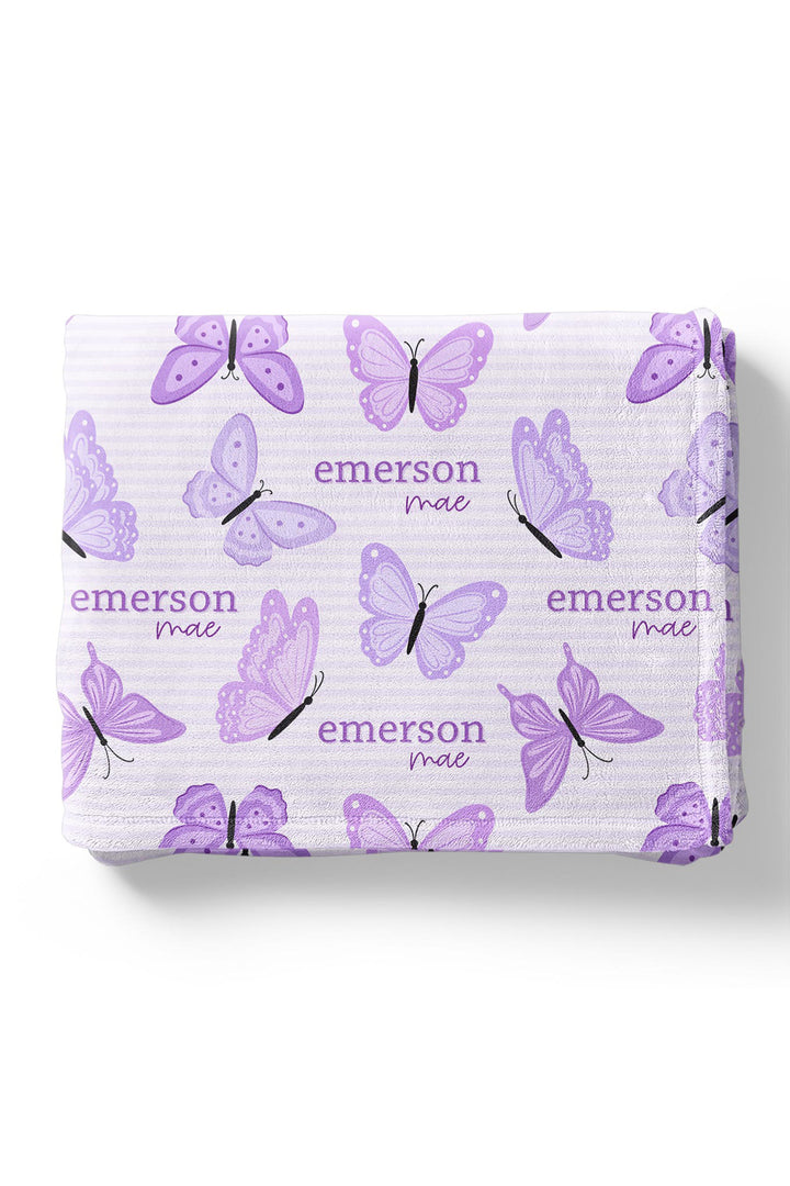 Flat lay of a folded personalized Caden Lane Lavender Wings blanket in lavender striped fabric, showing purple butterflies and custom name print