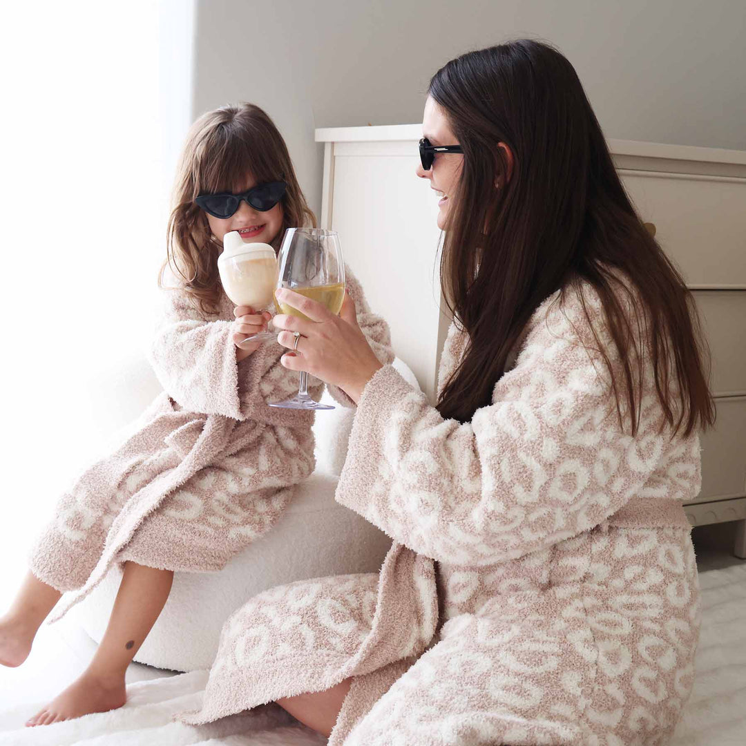 mom and daughter sitting and cheersing a wine glass and sippy cup in matching leopard robes 