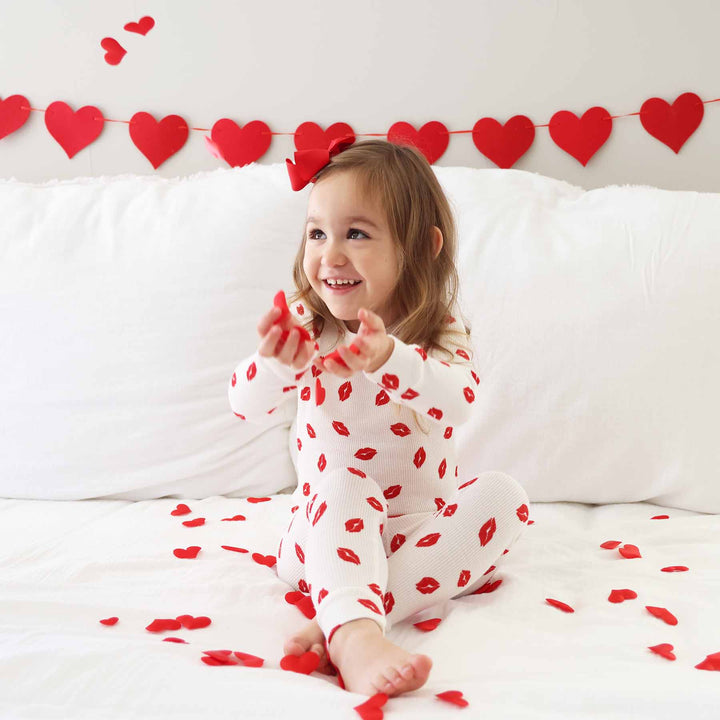 girl sitting on bed wearing a red lip valentine's day pajama set throwing heart confetti into the air 