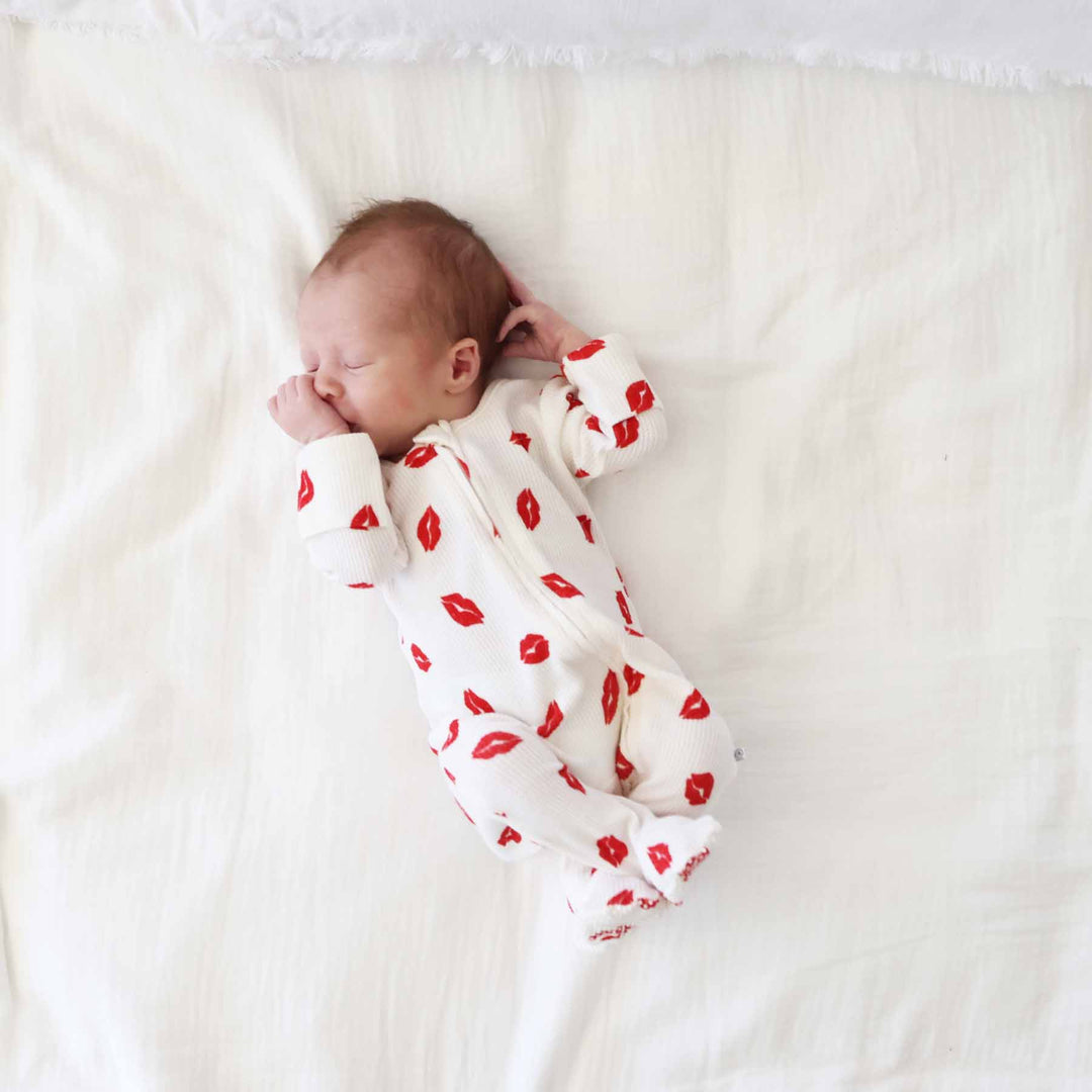 newborn boy laying on white blanket wearing a red lipstick kiss bamboo waffle zipper footie pajama for babies 