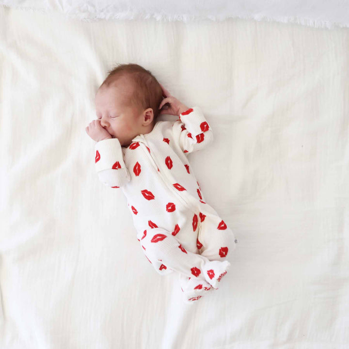 newborn boy laying on white blanket wearing a red lipstick kiss bamboo waffle zipper footie pajama for babies 