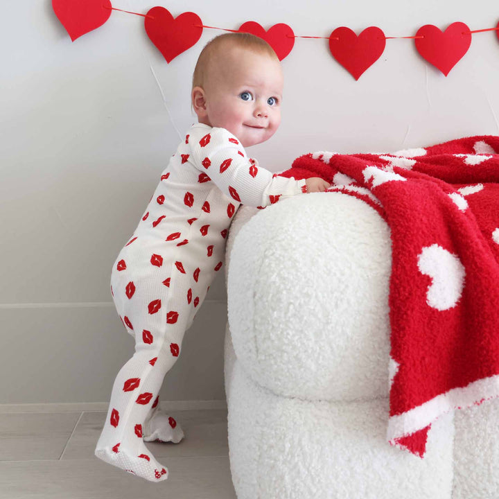 baby boy standing against an ottoman in a lipstick kisses zipper footie 