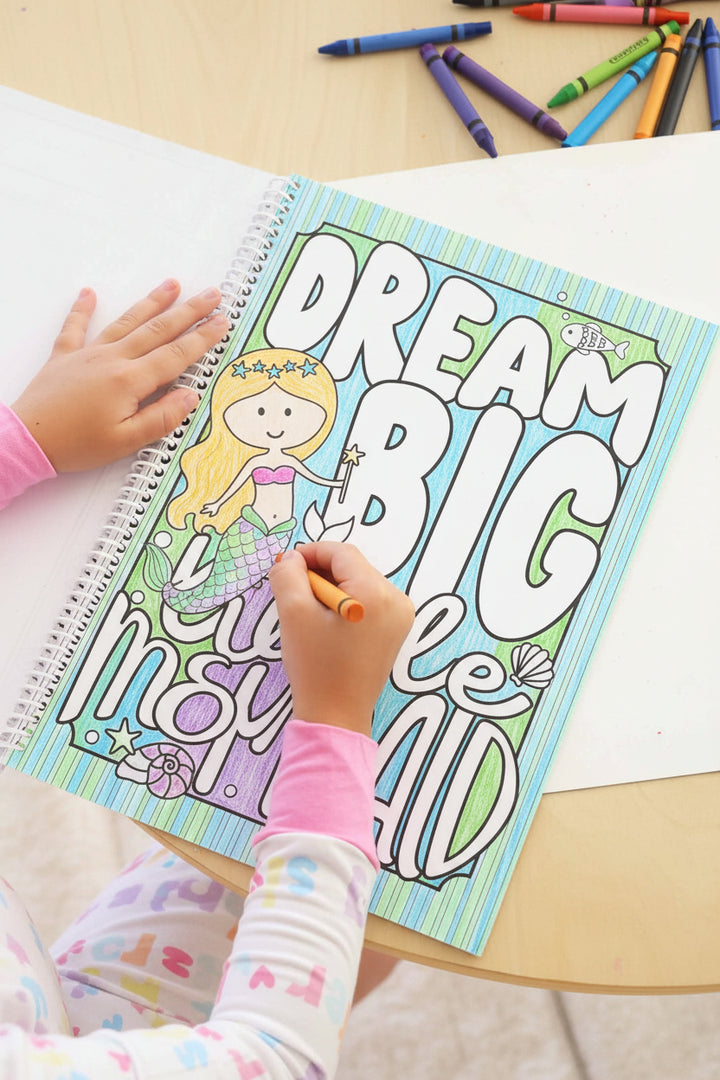 Child in a Big Sister pajama set coloring a 'Dream Big Little Mermaid' page with an orange crayon, filling in a mermaid illustration in a Caden Lane coloring book.