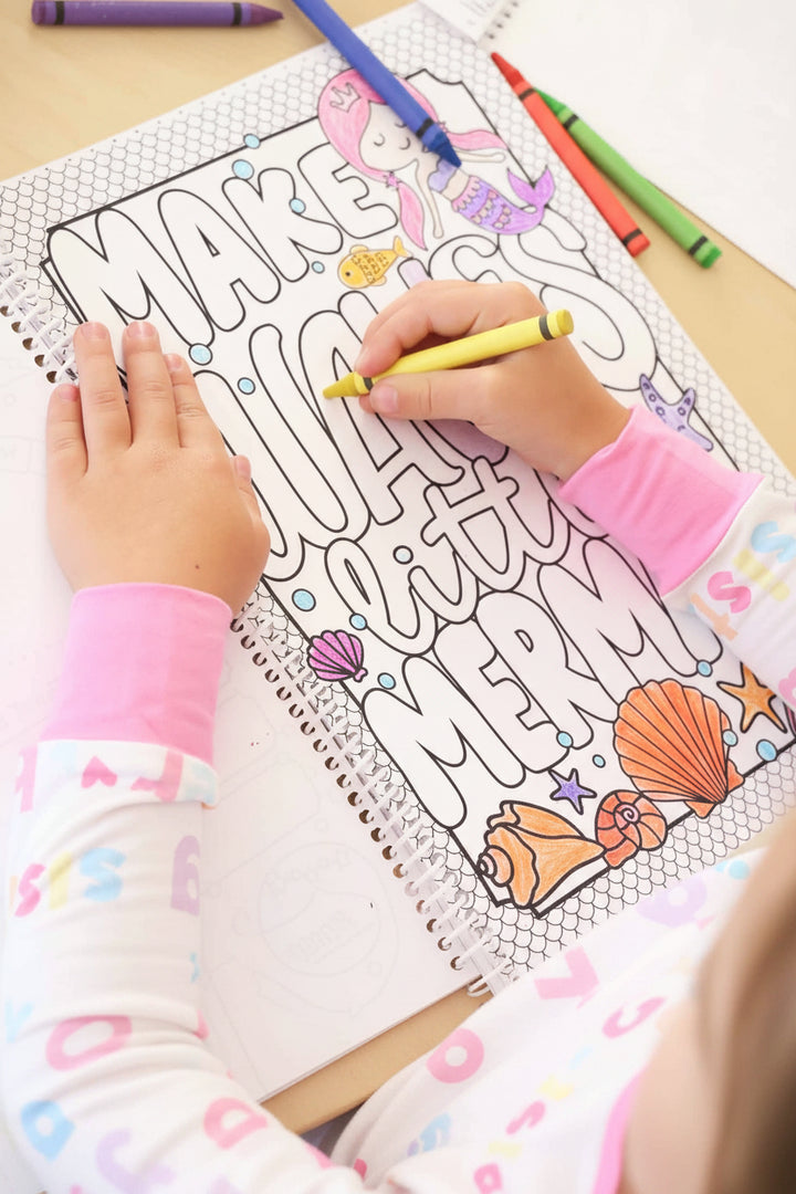 Child in a Big Sister pajama set coloring a 'Make Waves Little Mermaid' page with a yellow crayon in a Caden Lane coloring book, surrounded by seashell illustrations
