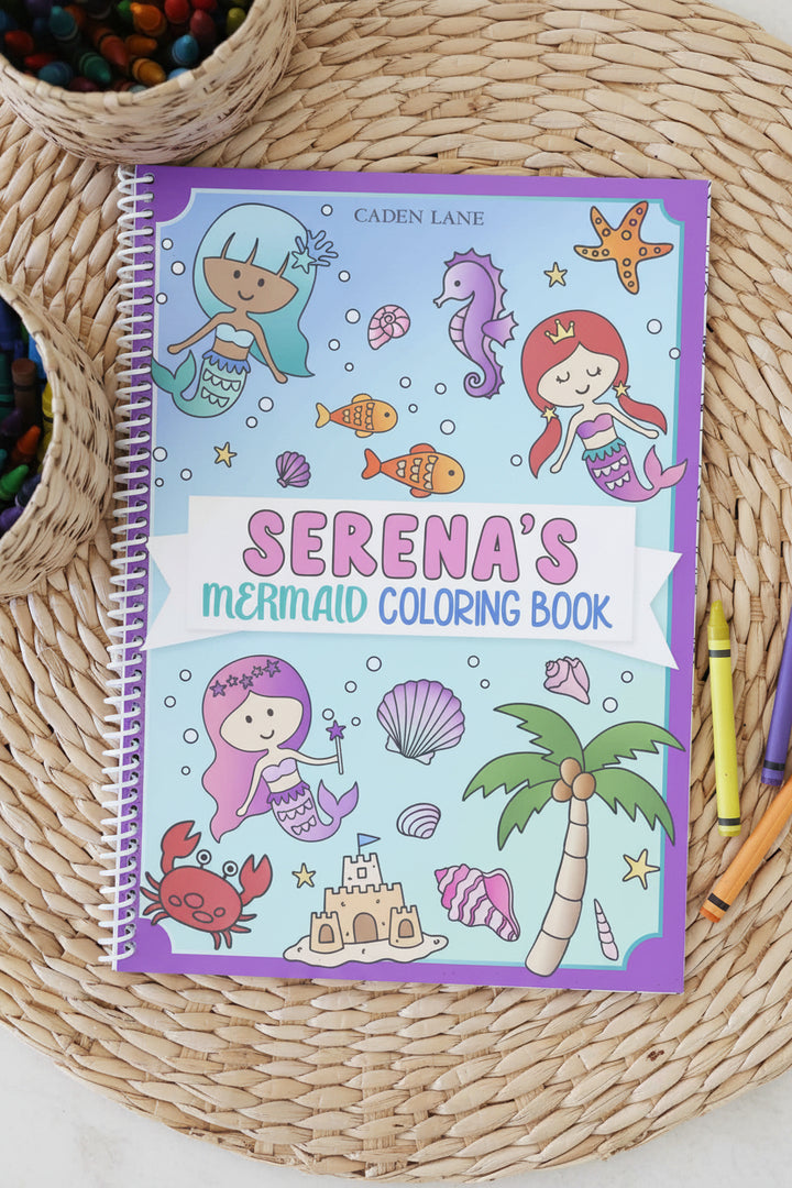 Caden Lane 'Serena's Mermaid Coloring Book' with an underwater cover featuring mermaids, seahorses, and a palm tree, displayed on a woven mat with a crayon basket