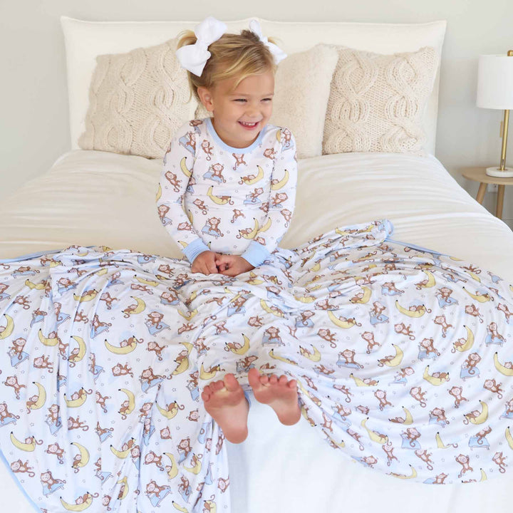 girl sitting on bed in monkey see, monkey snooze two piece pajama set with bamboo blanket draped over her 