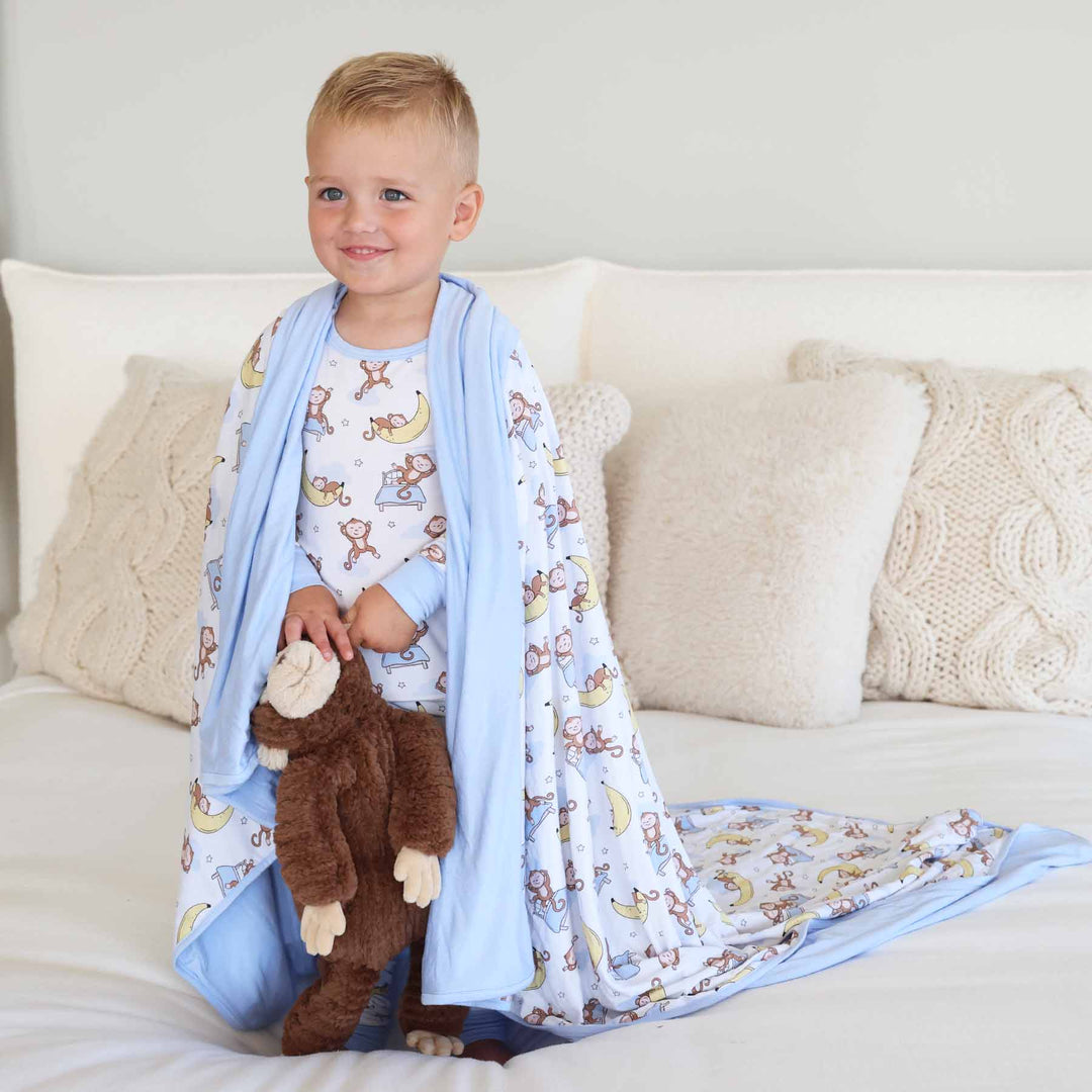 boy standing on bed wearing money and banana bamboo two piece pajama set with matching blanket draped over him 