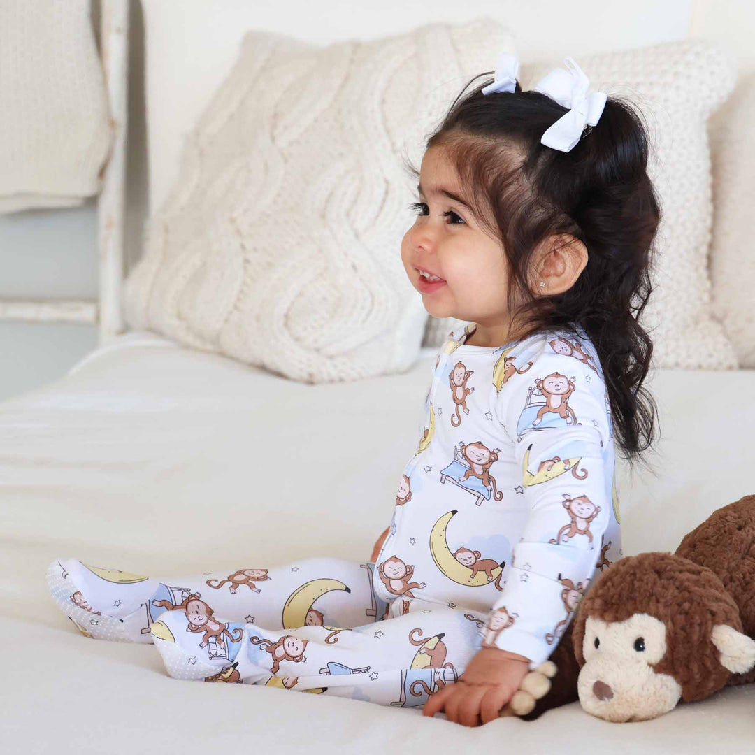 baby girl sitting on a bed wearing a monkey see, monkey snooze gender neutral baby footie with a monkey stuffed animal