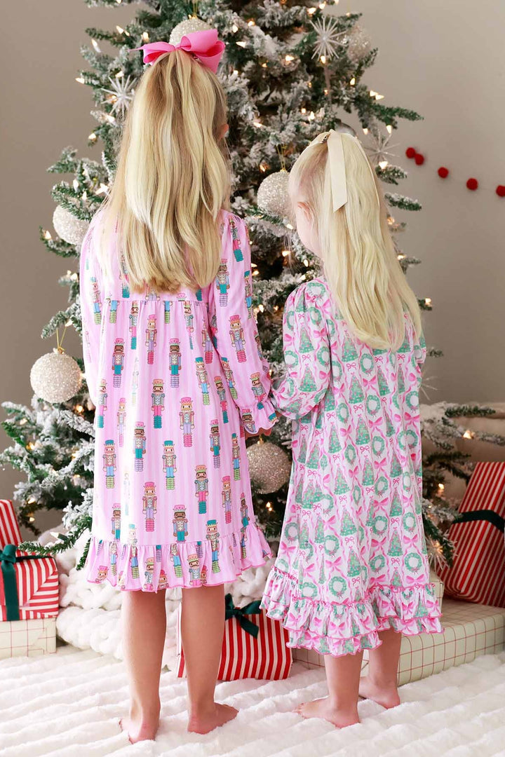 Two girls stand beside a Christmas tree, one wearing a pink Caden Lane nightdress with wreaths and bows, and the other wearing a pink nightdress featuring nutcracker prints