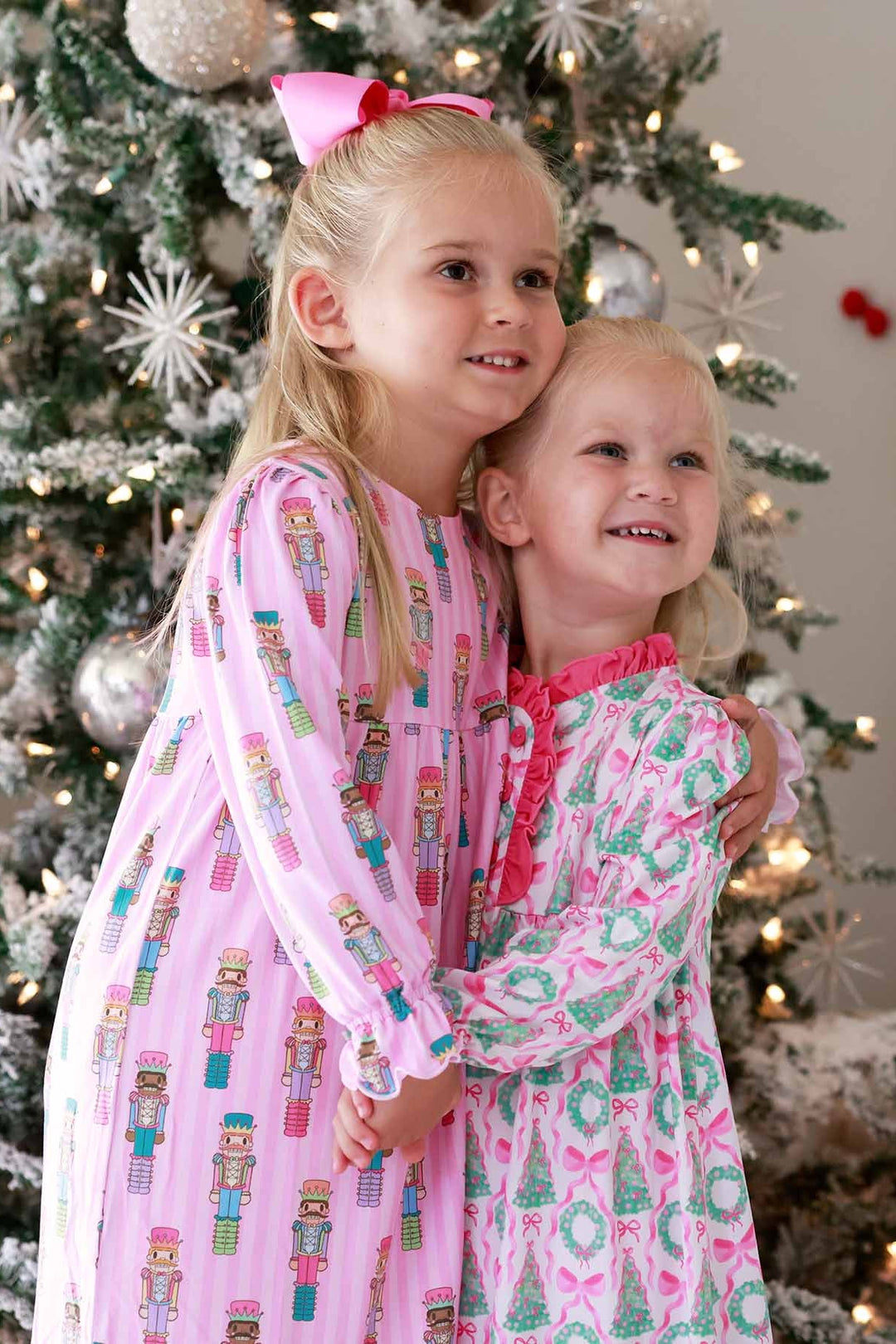 Two girls hugging beside a Christmas tree, one wearing a pink Caden Lane nightdress with wreaths and bows, and the other wearing a pink nightdress featuring nutcracker prints