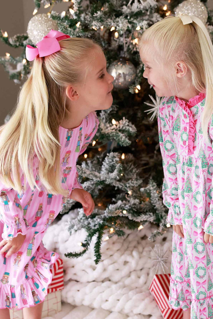 Two girls stand beside a Christmas tree, one wearing a pink Caden Lane nightdress with wreaths and bows, and the other wearing a pink nightdress featuring nutcracker prints