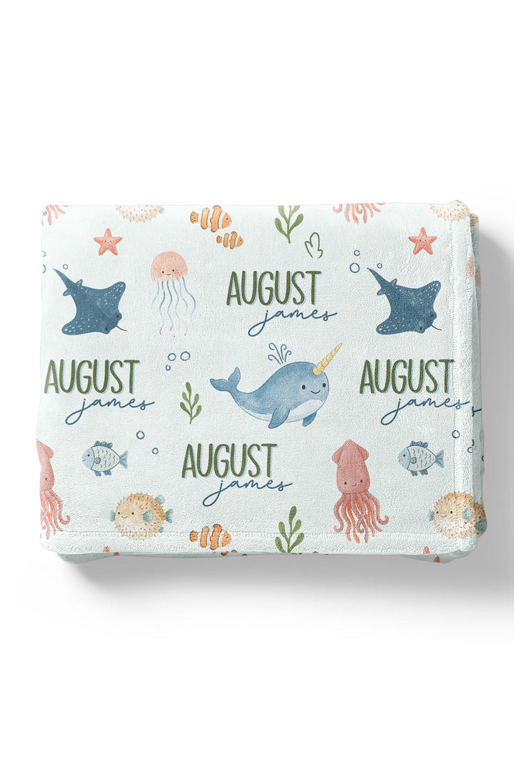 Flat lay of a folded personalized Caden Lane Ocean Dreams blanket in seafoam, showing narwhal, stingray, octopus, and custom name print