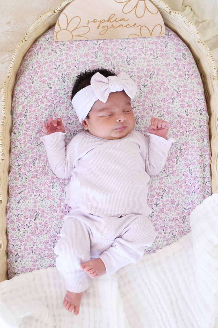 baby girl laying in basket in orchid two piece waffle crossover set with purple floral swaddle under her and a wood name sign 