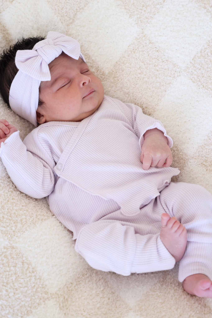 baby girl laying on neutral check blanket wearing a bamboo crossover set in pastel purple with a matching headwrap