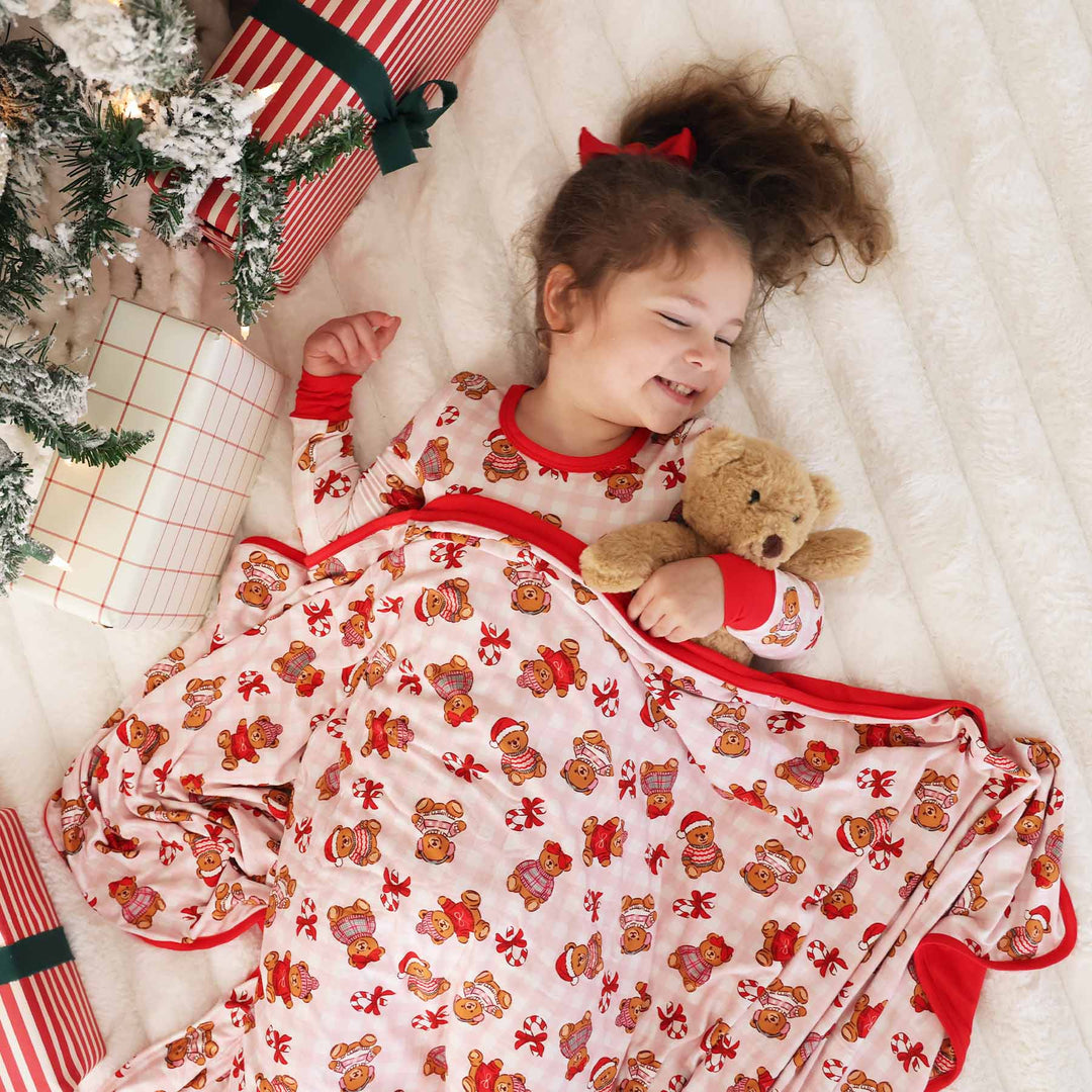 girl laying under the tree in a christmas pajama set with teddy bears wearing christmas sweaters holding a teddy bear with a matching blanket over her 