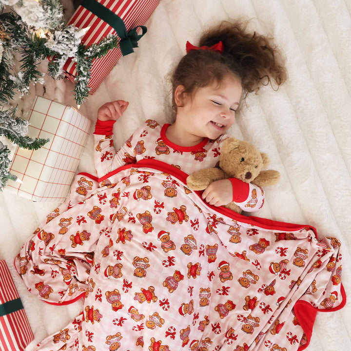 girl laying under the tree in a christmas pajama set with teddy bears wearing christmas sweaters holding a teddy bear with a matching blanket over her 