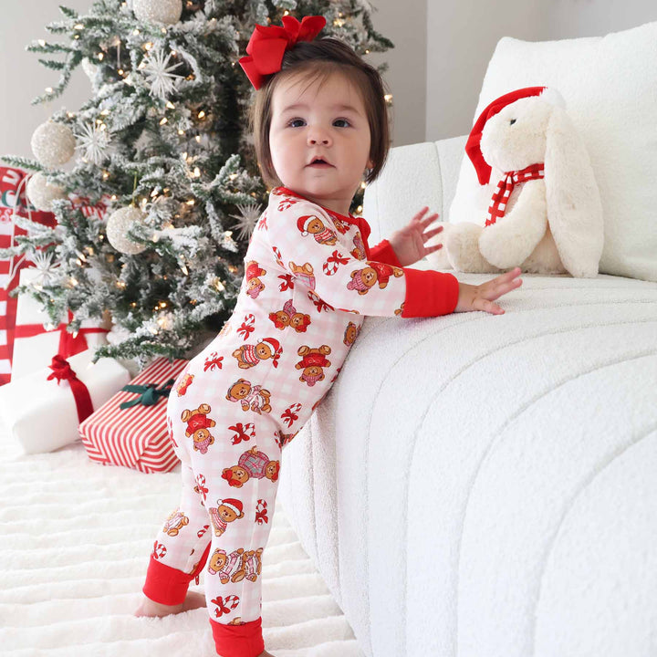toddler girl standing next to couch with pink gingham teddy bear pajama romper with a red bow in her hair 