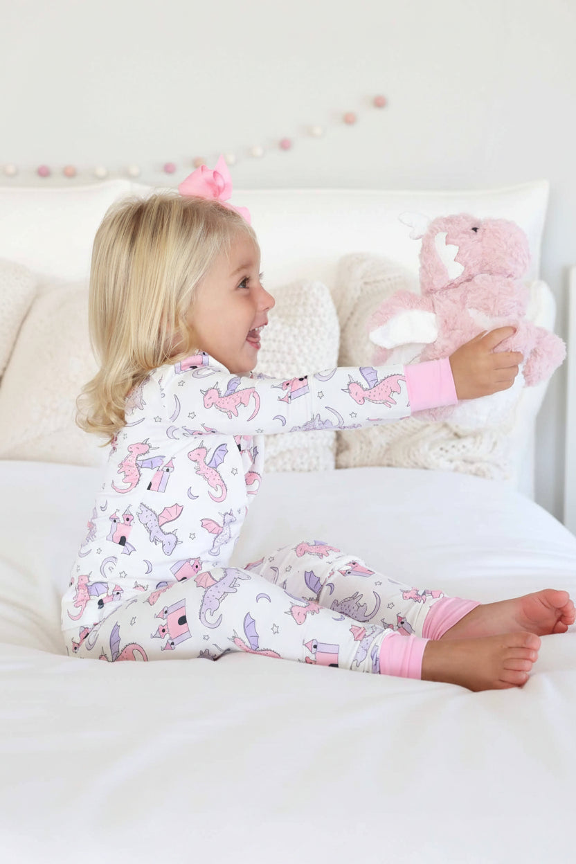 girl wearing pink and purple dragon and castle long sleeve and pants pajama set holding a pink dragon stuffed animal 