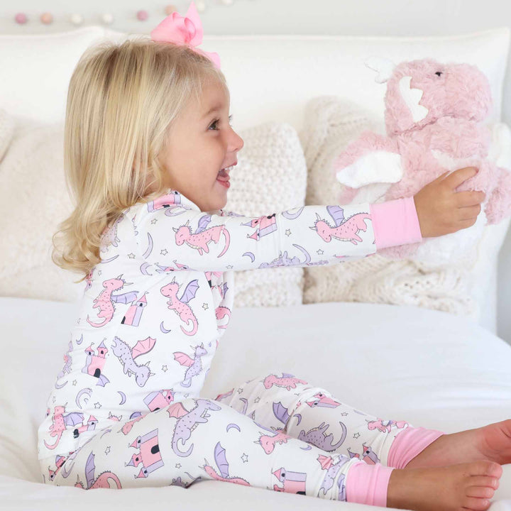 girl wearing pink and purple dragon and castle long sleeve and pants pajama set holding a pink dragon stuffed animal 