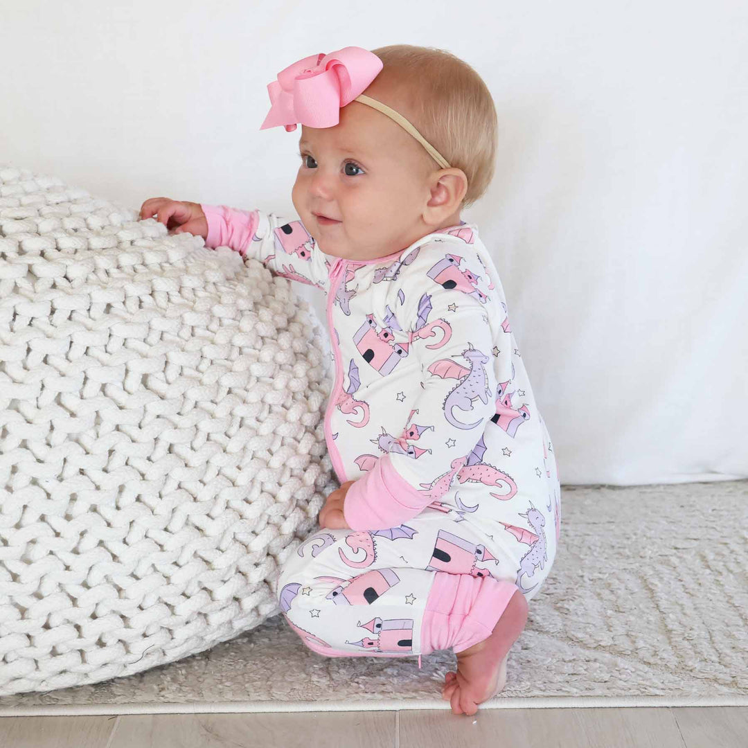 crouching baby girl in pink and purple dragon and castle pajama romper with matching solid pink ribbon bow headband 