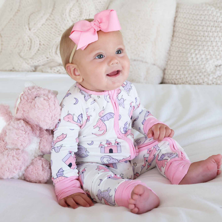 baby girl sitting on bed wearing pink dragon adventures convertible zip romper with matching ribbon bow headband with a pink dragon stuffed animal