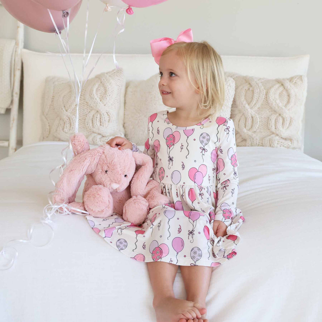 girl sitting on bed wearing pink balloon long sleeve dress next to a pink bunny stuffed animal