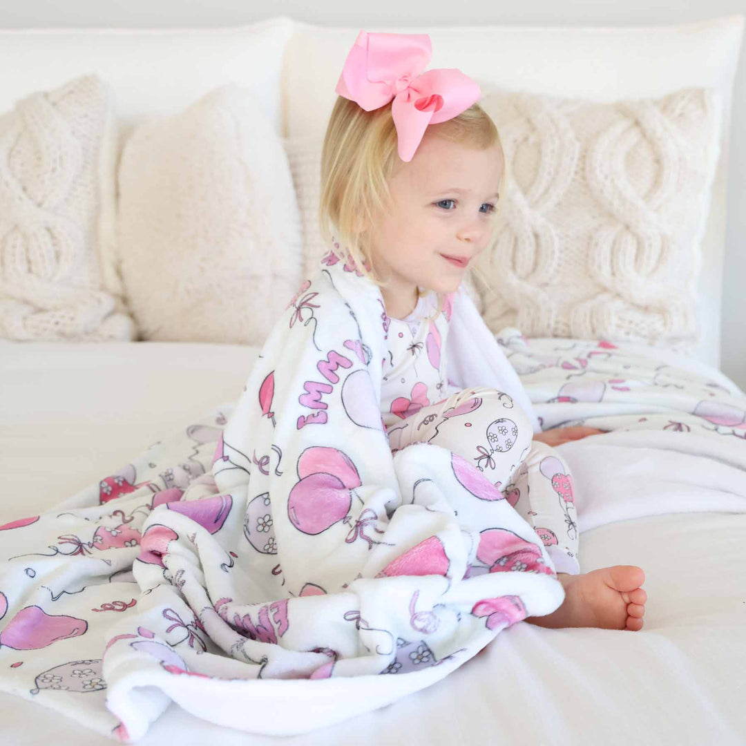 girl sitting on bed with personalized pink balloon and streamer blanket draped over her shoulders 
