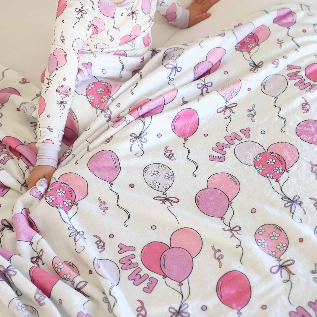 girl sitting on bed wearing a pink balloon long sleeve pajama set covered with a matching personalized blanket 