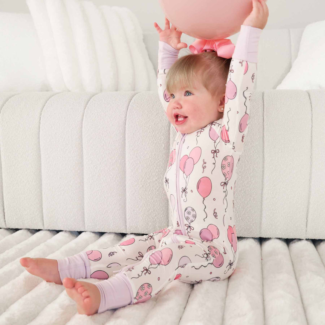girl wearing pink and purple balloon zip romper pajama holding a balloon above her head 