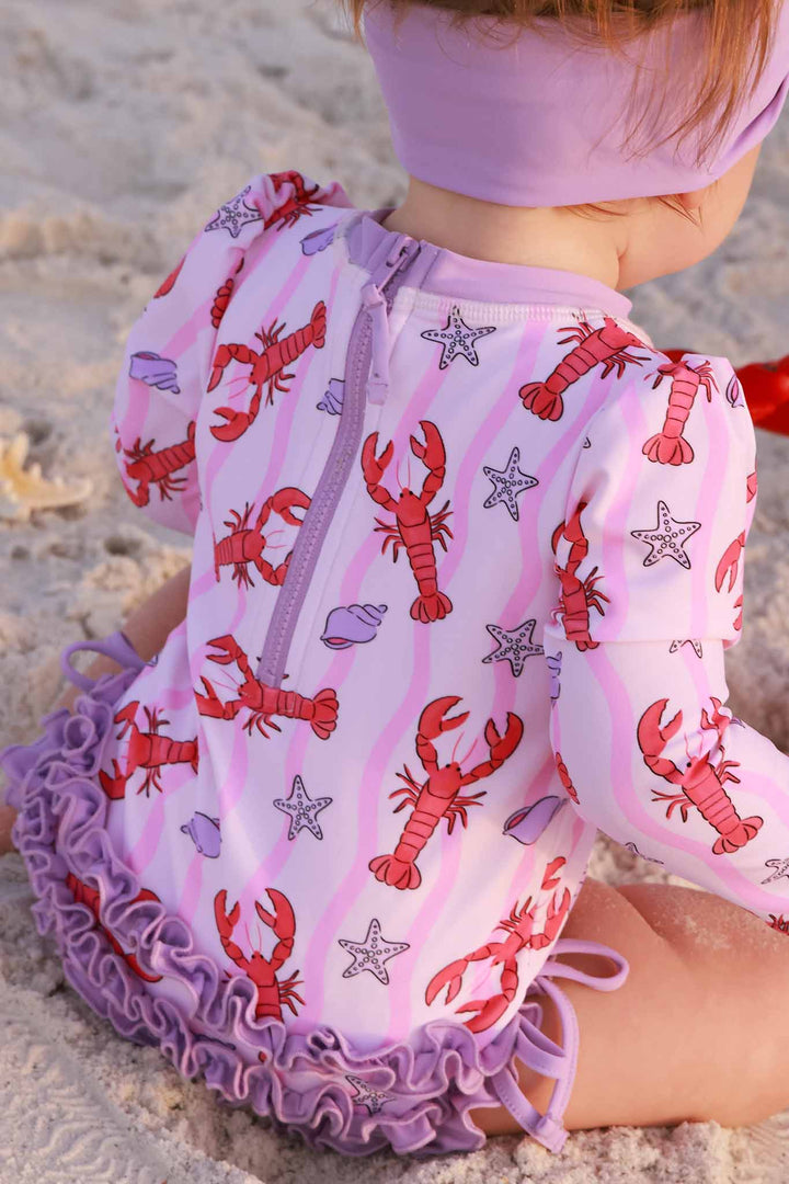 baby girl sitting on beach in long sleeve ruffle bottom rash guard swimsuit with lobsters
