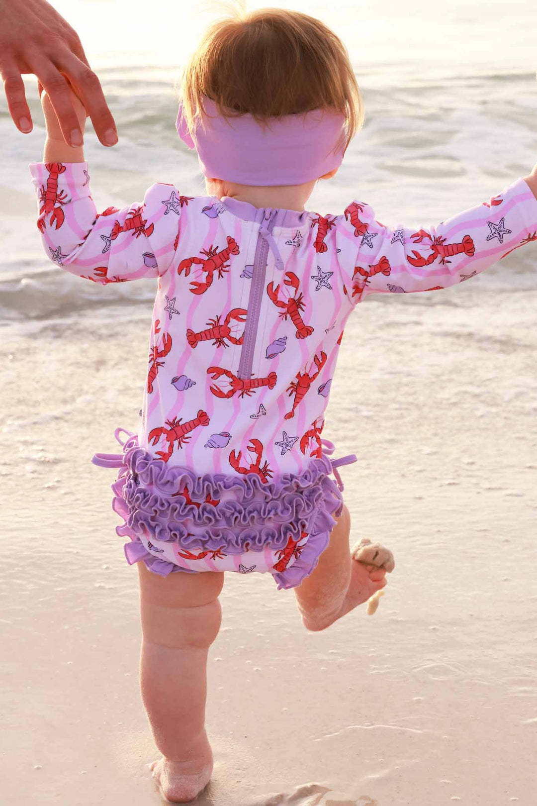 baby girl walking on the beach towards the ocean in pinch me ruffle bottom swimsuit 