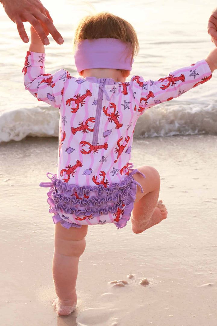 baby girl playing on the beach in lobster purple ruffle bottom long sleeve swimsuit 