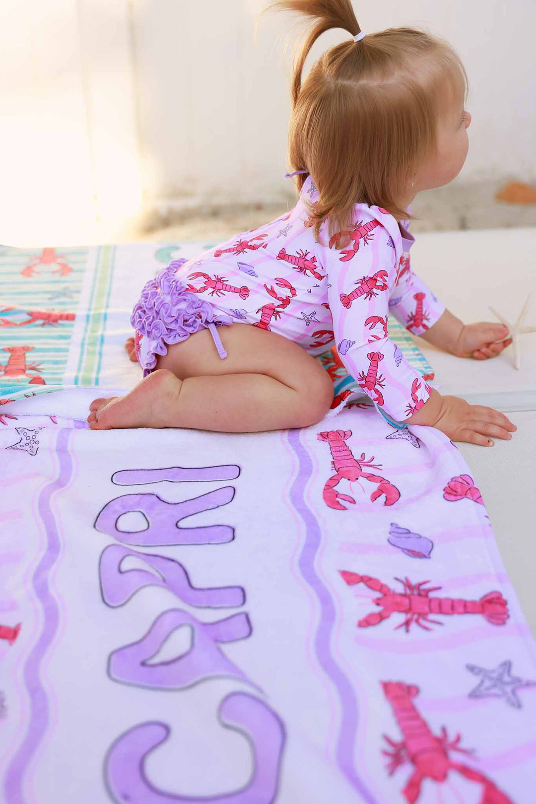 baby girl crawling on beach chair in pinch me pink long sleeve rash guard with ruffle bottom with a pony tail in her hair 