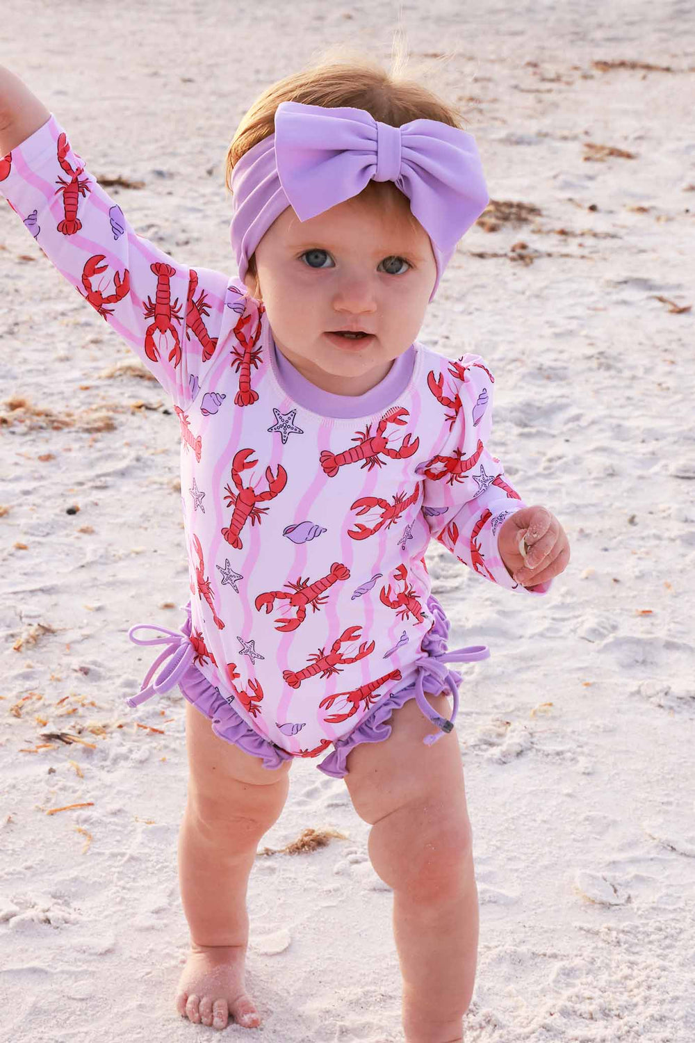 baby girl walking on the beach in long sleeve rash guard swimsuit with ruffle bottom in pinch me lobster print wearing a matching light pink swim bow 